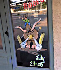 [A bull rider riding a snorting bull is painted on the window of a door of a business in town. The fair dates are in the lower right corner.]